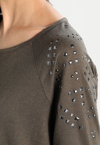 Olivgrüner Pullover mit einem offenen Ausschnitt, der mit metallischen Nieten und strukturierten Akzenten an den Schultern für zusätzliche Details versehen ist.