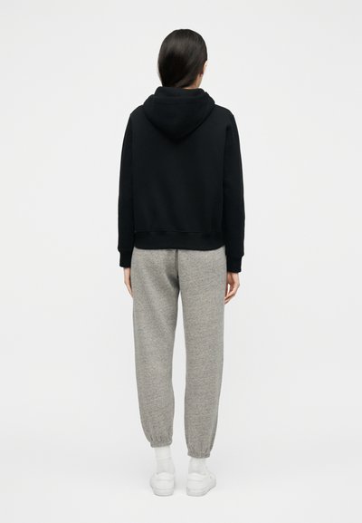 Γυναίκα με την πλάτη γυρισμένη, φοράει μαύρη φούτερ με κουκούλα, γκρι φόρμα, άσπρες κάλτσες και άσπρα αθλητικά παπούτσια μπροστά σε απλό λευκό φόντο.