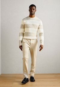 Kermainen raidallinen neule, jossa on teksturoitu kuvio, yhdistetty beigen värisiin housuihin. Mustat kengät täydentävät asua. Malli seisoo neutraalin taustan edessä.