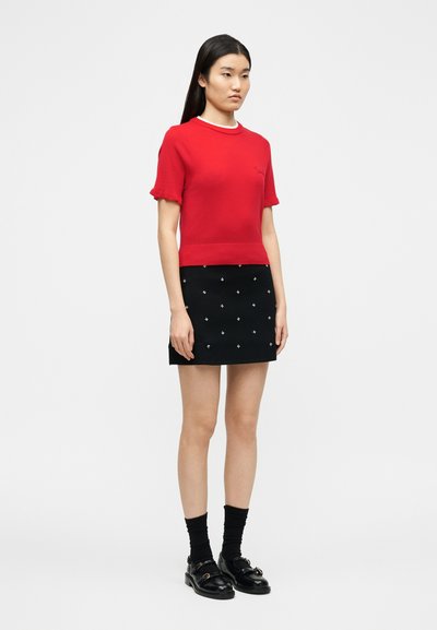 Νεαρή γυναίκα στέκεται στραμμένη προς τα αριστερά, φορώντας κόκκινη μπλούζα με κοντά μανίκια, μαύρη μίνι φούστα με λευκά σταυρωτά σχέδια, μαύρες κάλτσες και μαύρα μοκασίνια.