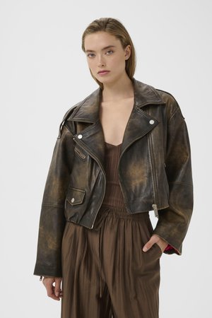 Młoda kobieta ubrana w wyciąganą czarną skórzaną kurtkę motocyklową na brązowym prążkowanym topie i luźnych brązowych spodniach, stojąca na jednolitym tle.