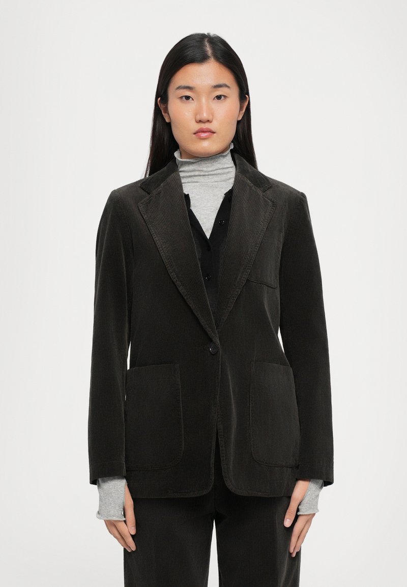 Dark green corduroy blazer with notched lapels, two front pockets, single button closure, worn over a gray turtleneck.