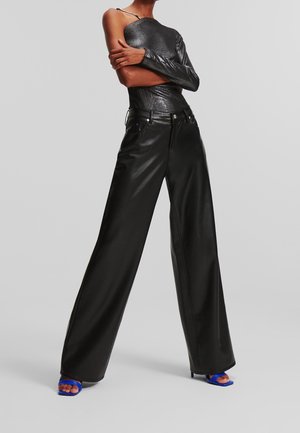 Persoon draagt een zwarte top met één schouder, zwarte leren broek met wijde pijpen en blauwe open-toe sandalen met hak, poserend tegen een grijze achtergrond.