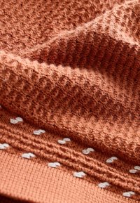 Strukturierter Strickstoff in Rostfarbe mit einem Muster aus kleinen weißen Akzenten; der Stoff ist weich und leicht flauschig.