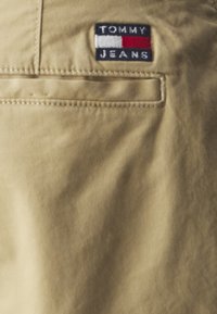 Tela beige con un bolsillo trasero que cuenta con un pequeño logotipo bordado en rojo, blanco y azul, que dice "TOMMY JEANS". Textura suave.
