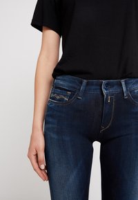 Jeans de mezclilla azul oscuro con un diseño ajustado, con costuras claras, un pequeño bolsillo trasero con estampado y un cierre frontal estándar con botón.