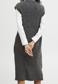 Robe en maille côtelée grise avec une fente latérale, à manches courtes, portée sur une chemise blanche à manches longues. Tissu texturé.