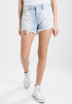 Personne portant un short en jean déchiré bleu clair et des baskets montantes blanches, debout devant un fond uni et clair.