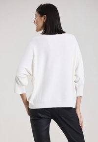 Weißer Strickpullover mit entspannter Passform, Bootsausschnitt und Dreiviertelärmeln. Kombiniert mit schwarzen Hosen, die eine glatte Textur und klare Linien zeigen.