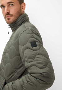 Polstret quiltet jakke i olivengrøn med hexagonmønster, høj krave og et sort stofmærke på skulderen. Lynlås foran.