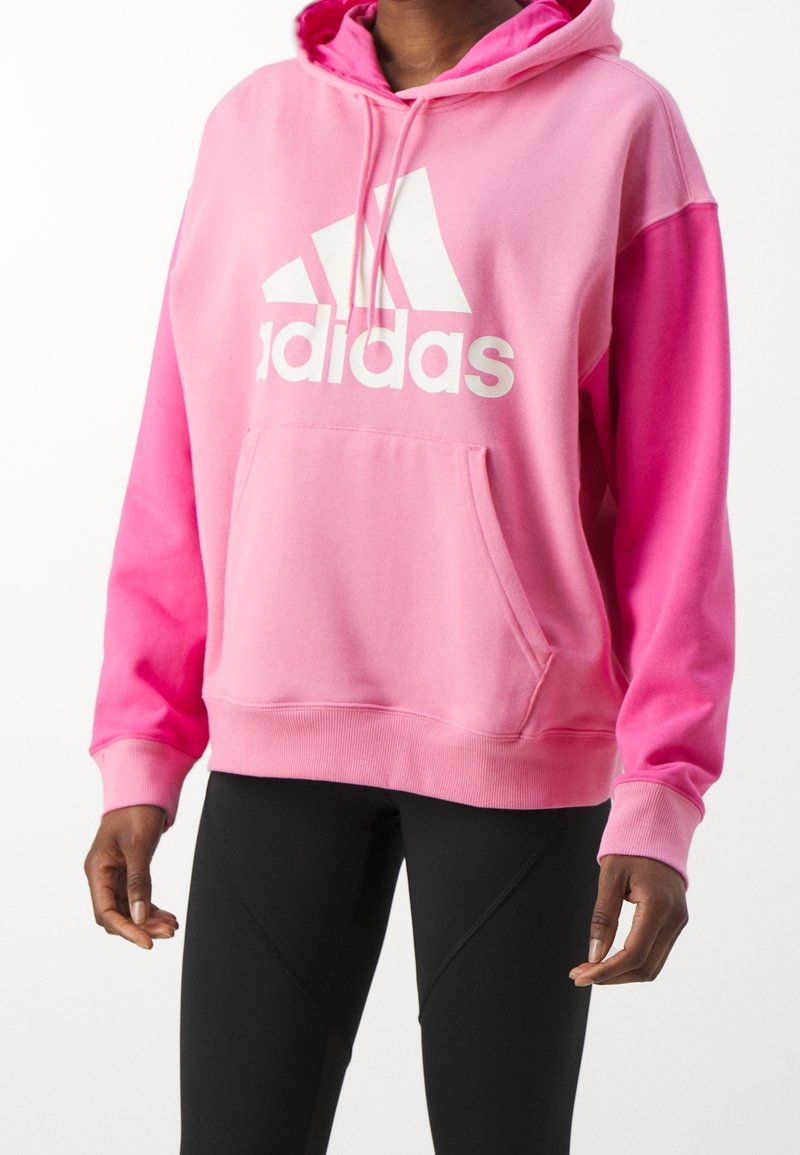 Rosa huvtröja med en stor vit Adidas-logotyp, framficka, ribbade ärmslut och en avslappnad passform. Tillverkad av mjukt tyg.