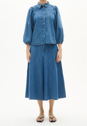 Kobieta ubrana w niebieską denimową bluzkę na guziki z bufkami, zestawioną z szerokimi spodniami culottes z denimu i czarnymi klapkami.
