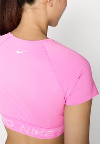 Roza športna crop majica iz gladkega materiala, s kratkimi rokavi in rebrastim pasom, na katerem je spredaj natisnjen logotip "Nike" v beli barvi.
