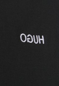 Tissu noir avec texte brodé blanc "HUGO" en miroir horizontal.