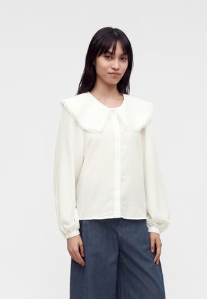 Jonge vrouw met lang donker haar, die een witte blouse draagt met een grote kanten kraag en losse mouwen, gecombineerd met brede blauwe broek.