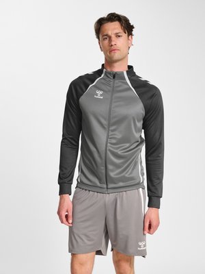 Homme portant une veste de sport zippée grise et noire et un short de sport gris, debout devant un fond blanc uni.