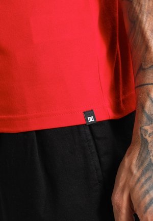 Ourlet de chemise rouge avec une petite étiquette noire du logo DC, portée sur un pantalon noir, et un avant-bras tatoué reposant à côté des vêtements.