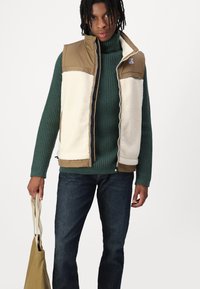 Gilet foderato in pile, colori beige e crema, chiusura con zip, colletto alto, indossato sopra un maglione a coste verde scuro, abbinato a jeans scuri e una borsa in tela.