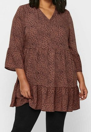Tunique marron ample avec motif tacheté noir, col en V, manches trois-quarts évasées et ourlet à volants portée avec un pantalon noir.