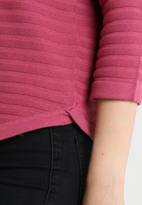 Różowy sweter w prążki wykonany z teksturowanej przędzy, z lekko rozszerzanym dołem i podwiniętymi mankietami, noszony na czarnych dżinsach.