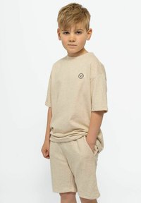 Beige puuvillane t-särk ja sobivad lühikesed püksid. T-särgil on lühikesed varrukad ja ümmargune kaelus, millel on rinnal väike logo.