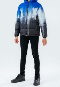 Giacca imbottita blu e nera con un motivo a goccia in stile graffiti, dotata di chiusura con zip, cappuccio e polsini elasticizzati, abbinata a pantaloni neri e sneakers.