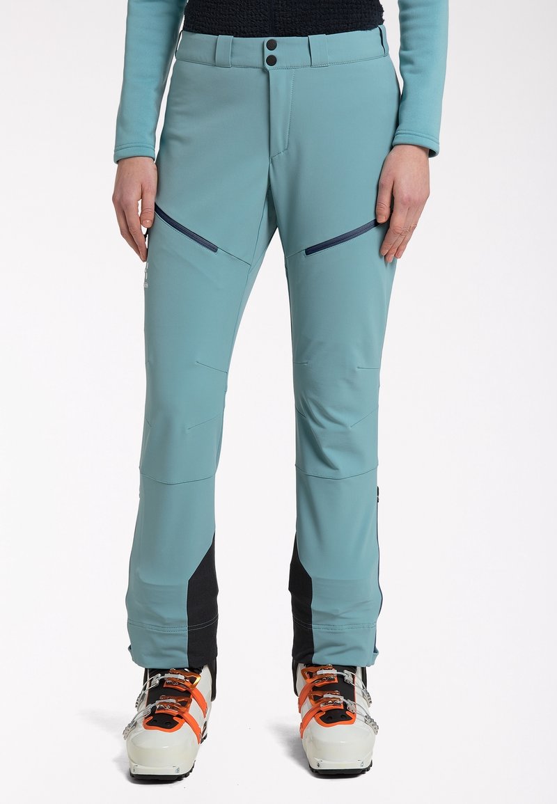 Personne portant un pantalon de ski bleu sarcelle avec des empiècements noirs et des chaussures de ski blanches avec des sangles orange, debout devant un fond blanc uni.