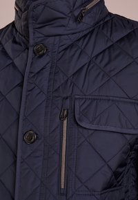 Granatowa pikowana kurtka z wysokim kołnierzem, z przodu na guzik oraz z kieszenią na zamek. Właściwości obejmują diamentowy wzór i gładką teksturę.