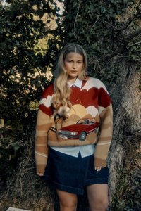 Strickpullover mit roten, beigen und braunen Wellen, der ein gesticktes rotes Auto zeigt. Kombiniert mit einem Jeansrock und einem darunter getragenen Hemd mit Kragen.