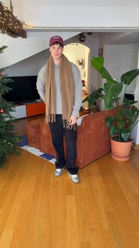 Ruskea huivi, harmaa pitkähihainen paita, mustat housut, hopeanväriset lenkkarit. Sisätilassa, taustalla kukkaruukku ja sohva.