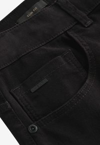 Slim fit black trousers with a smooth texture, featuring a small front pocket and a black branded tag. Subtle stitching details present.