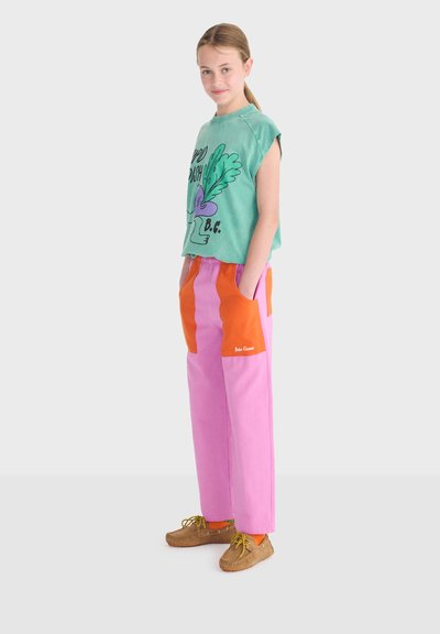 Jeune personne debout, les mains dans les poches d’un pantalon rose vif et orange, portant un haut sans manches vert et des mocassins couleur beige.