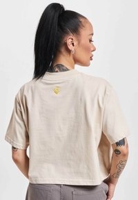 Beige cropped t-shirt with short sleeves, featuring a gold embroidery detail on the back. Made from soft fabric, showing subtle texture.