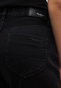 Nahaufnahme einer schwarzen Denim-Jeanstasche mit Pepe Jeans Logoaufnäher und eingesticktem Markennamen auf der Gesäßtasche.