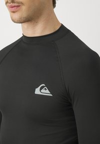 Svart rash guard laget av glatt, elastisk materiale. Har høy krage og en hvit logo på brystet. Tettsittende design.