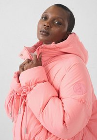 Giacca imbottita rosa con collo alto, dettagli con coulisse e finiture argento; presenta un logo circolare sulla manica. Materiale soffice e isolante.