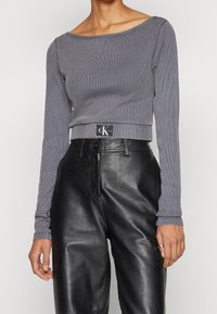 Grå ribbad långärmad crop top med vid halsringning, kombinerad med högmidjade svarta läderbyxor. Logotyp-patch synlig vid midjan.