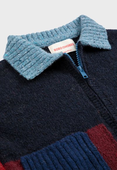 Kragpullover mit Reißverschluss, dunkelblauem Körper, roten und blauen Aufnähern sowie einem strukturierten hellblauen Kragen. Markenlabel sichtbar.