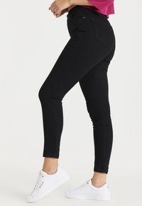 Vaqueros negros ajustados con una textura suave, cintura alta y diseño tradicional de cinco bolsillos. Combinados con zapatillas blancas con detalles dorados.