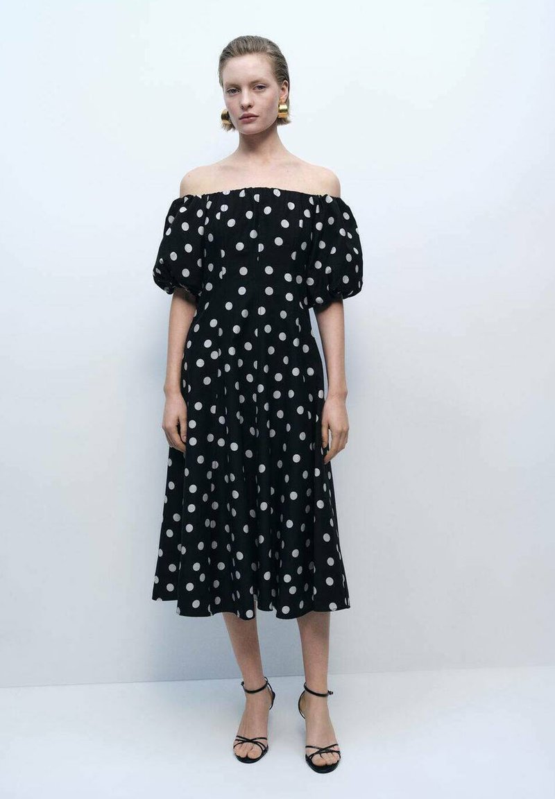 Rochie neagră cu umerii la vedere, cu buline albe mari, mâneci bufante, fustă fluidă și un material texturat. Sandale simple negre cu bretele.