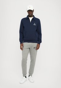 Sweatshirt azul-marinho com fecho e bolso frontal, calças de fato cinzas com o texto "POLO" e ténis brancos. Material em mistura de algodão, ajuste descontraído.