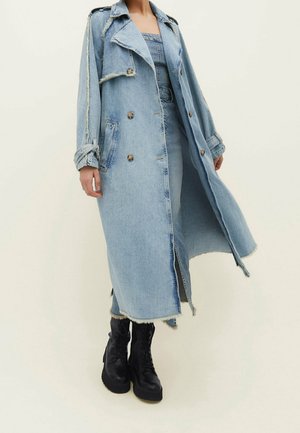 Osoba nosząca długi jasnoniebieski denimowy trencz z postrzępionymi krawędziami, dwurzędowymi guzikami i czarnymi butami na sznurowadła.