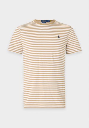 Ung mand iført en beige og hvid stribet t-shirt, tanfarvede knælange shorts, hvide sokker, hvide sneakers og en hvid kasket, stående med hænderne i lommerne.