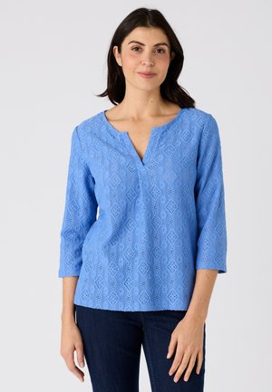 Top azzurro chiaro con motivo in pizzo, maniche a 3/4 e scollo a V con intaglio, indossato con denim scuro. La texture è intricata con motivi a diamante.