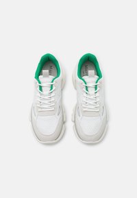 Białe buty sportowe z siateczkową cholewką, zielonymi akcentami wokół kołnierza i masywną podeszwą. Posiadają sznurówki oraz subtelne logo na języku.