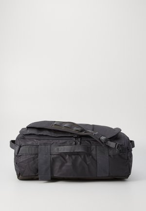 Czarna torba sportowa z miękkiego materiału z podwójnymi uchwytami, regulowanym paskiem na ramię i zapinanym na suwak głównym przegrodą, umieszczona na jednolitym jasnym tle.