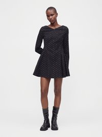 Femme portant une mini-robe noire à manches longues avec un petit motif coloré et des bottes noires mi-mollet, debout devant un fond blanc.