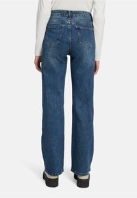 Jean bleu foncé taille haute avec une coupe décontractée, dotée de deux poches arrière et d'une texture en denim lisse. Associé à des chaussures à plateforme noires.
