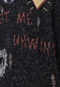 Črno pleteno blago z obrabljenimi luknjami in zbledelim rdečim ter belim vezenim napisom "PUSTI ME NOTRI, ZMAGUJEM".