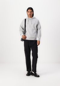 Šedá mikina s kapucí a přední kapsou, doplněná černými cargo kalhotami. Model nese malou černou crossbody tašku. Podlaha je bílá.